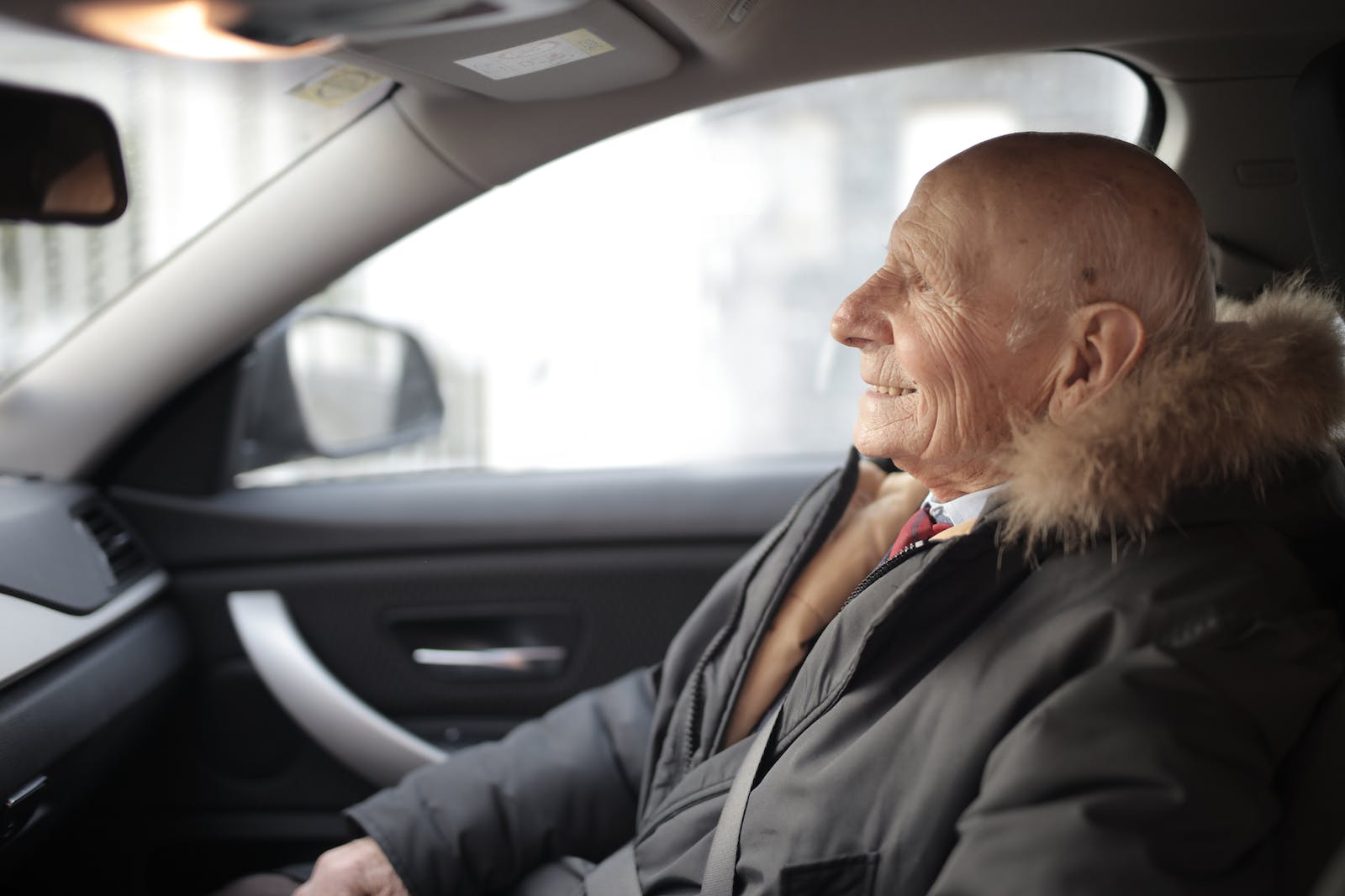 Side view of content elderly male in suit and outerwear sitting in front seat of contemporary automobile and looking away